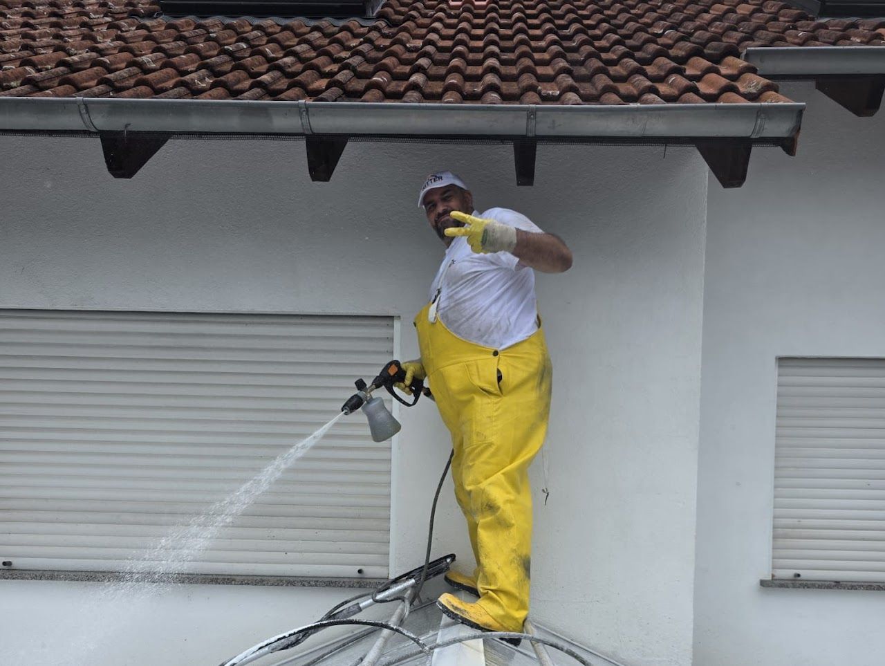 Eine Person auf einer Leiter reinigt ein weißes Gebäude mit einem Hochdruckreiniger. Sie trägt gelbe Schutzkleidung.