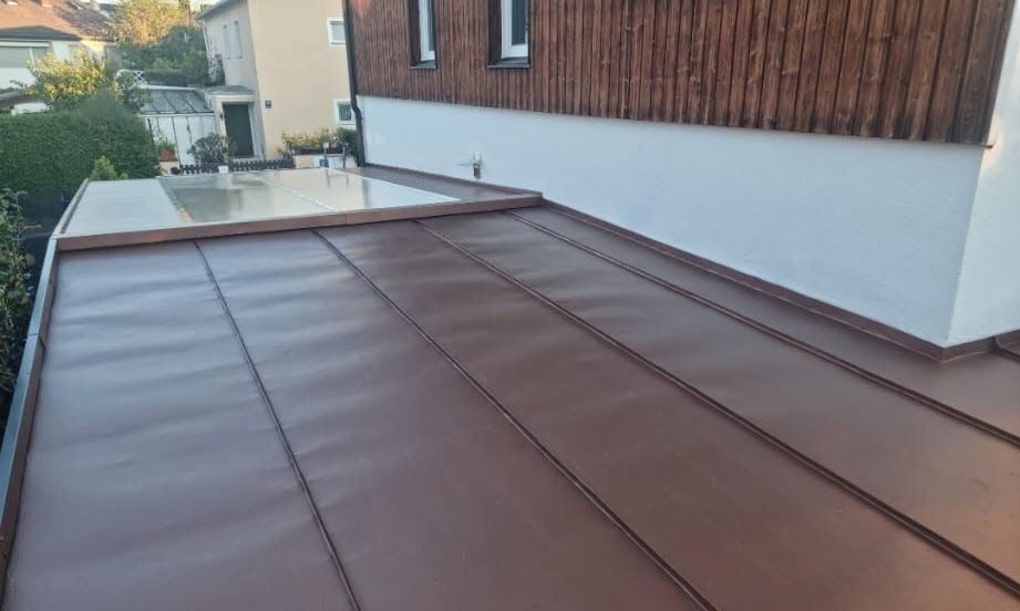 Braunes Metalldach eines Gebäudes mit einer Glaskonstruktion und einer weißen, holzgetäfelten Wand.