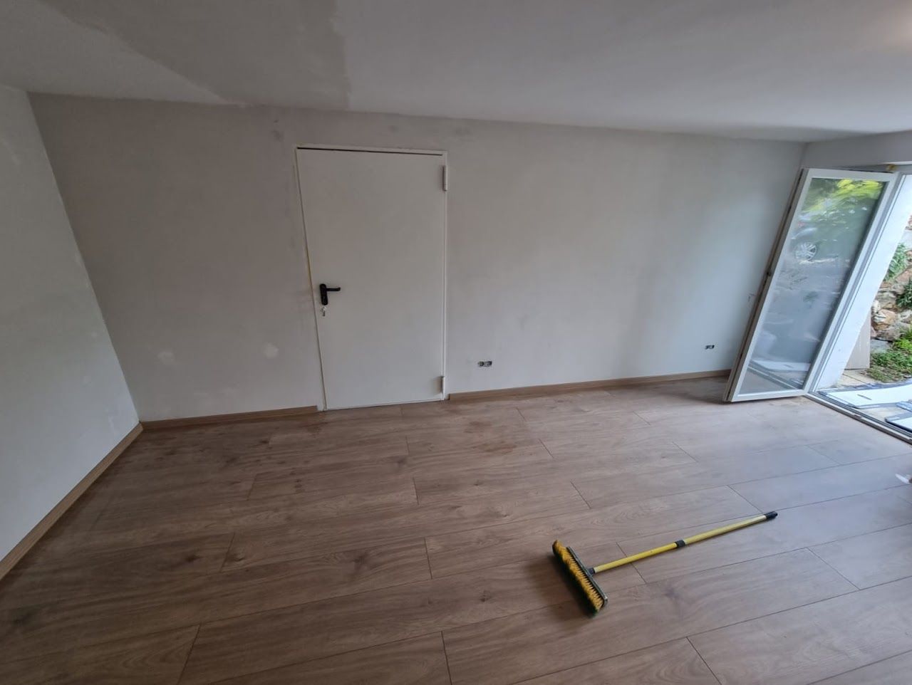 Leerer Raum mit Holzboden, weißen Wänden, geschlossener Tür und einem Besen.
