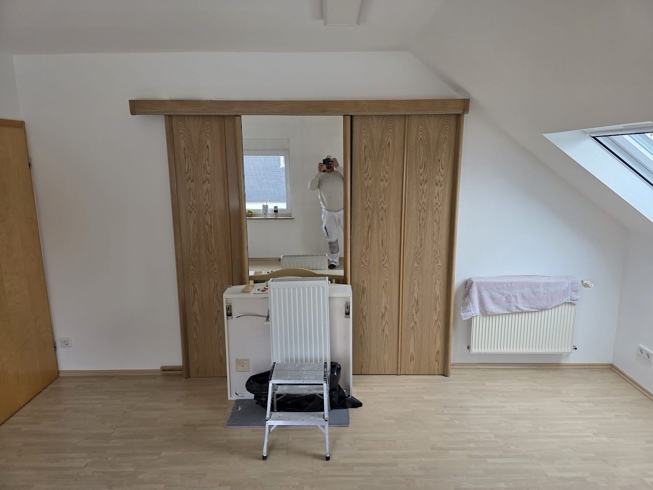Zimmer mit hölzernen Schiebetüren und einer Leiter, weißen Wänden und einem Heizkörper unter einem Fenster.