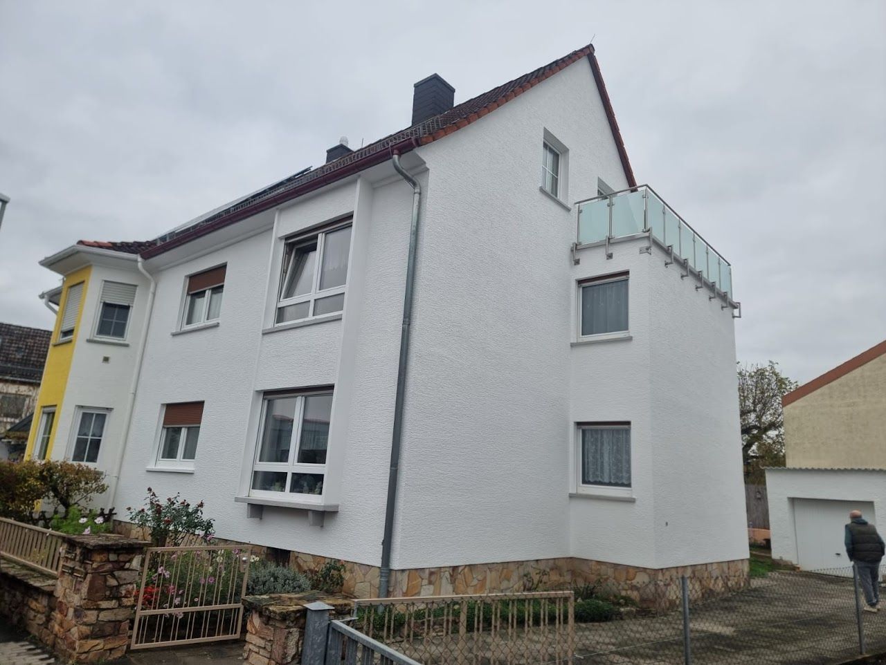 Weißes Stuckgebäude mit rotem Dach, Balkon und einer Person, die in der Nähe vorbeigeht.