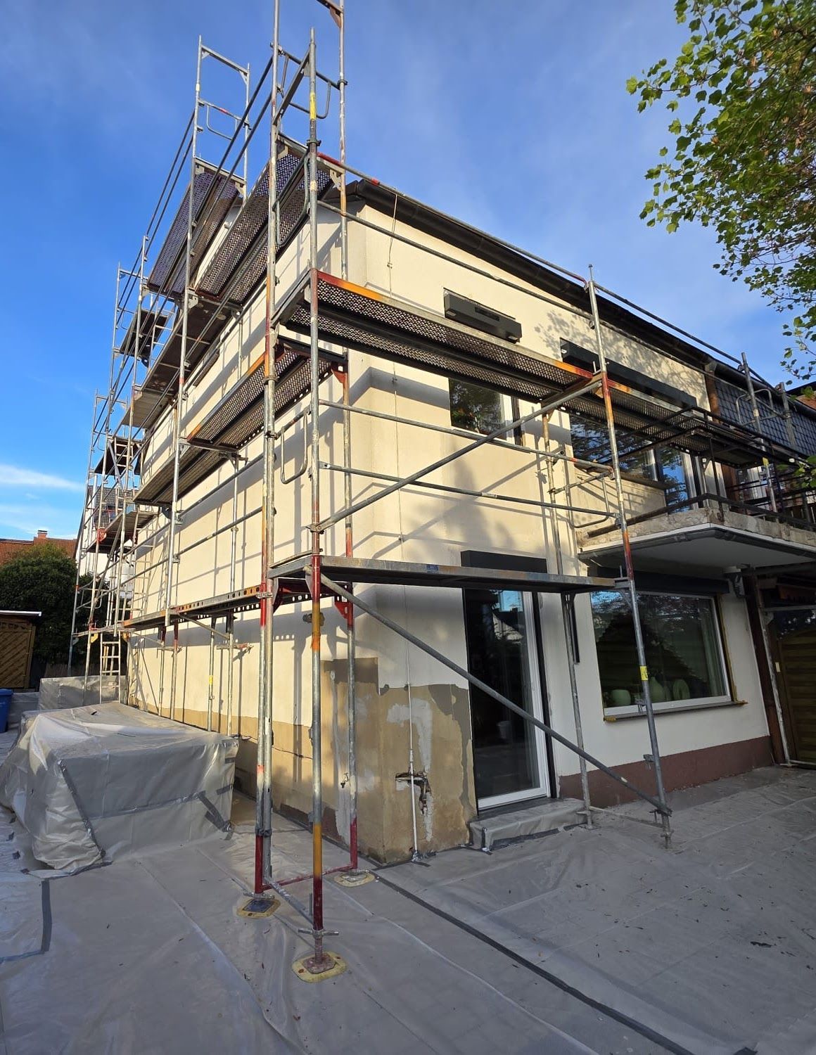 Das Gebäude befindet sich im Bau, ein Gerüst ist aufgebaut. Beige-weiße Fassade, der Boden ist mit einer Plane abgedeckt.