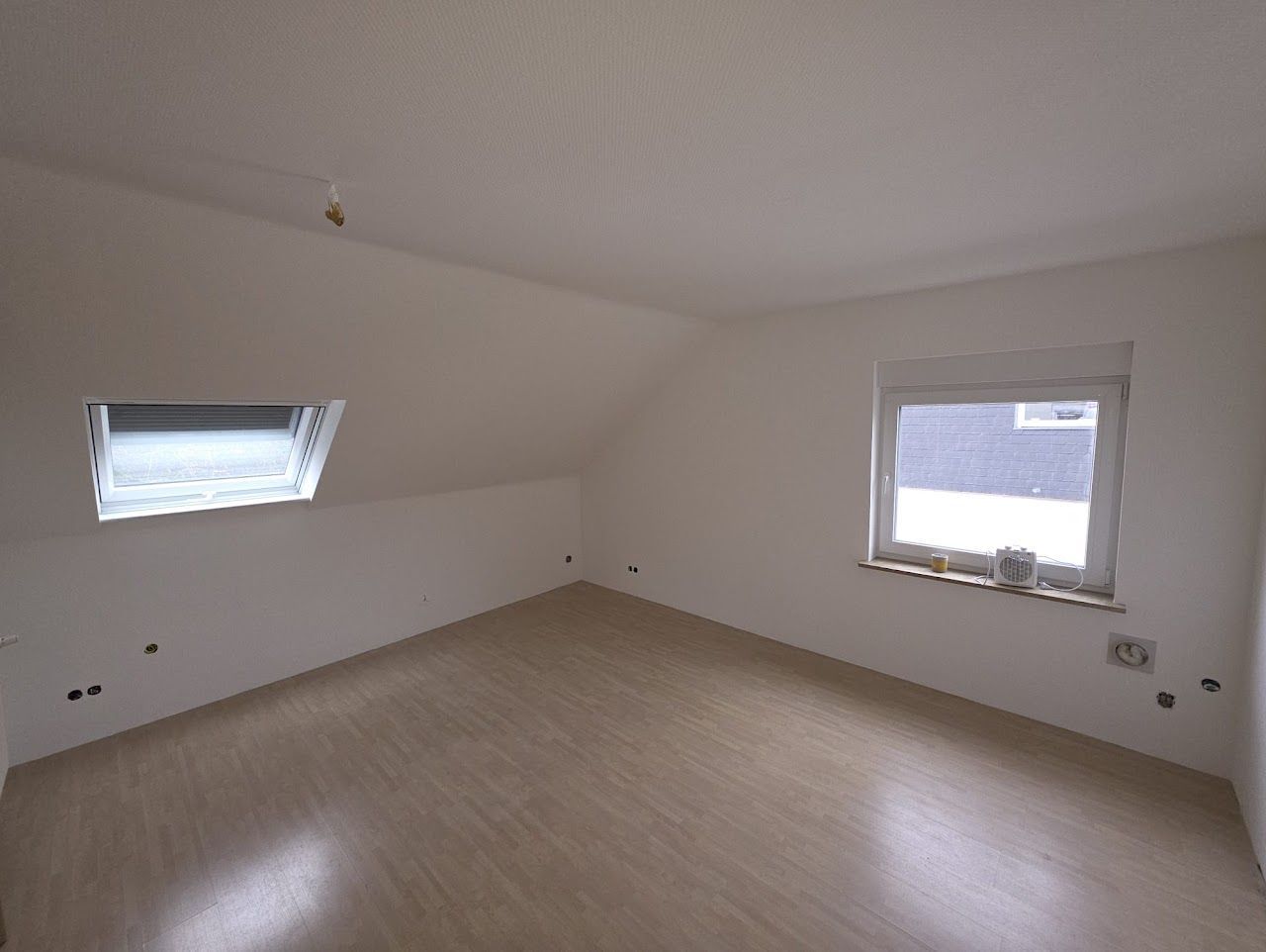 Leeres Zimmer mit Dachschräge, zwei Fenstern und Holzbodenoptik. Weiße Wände.