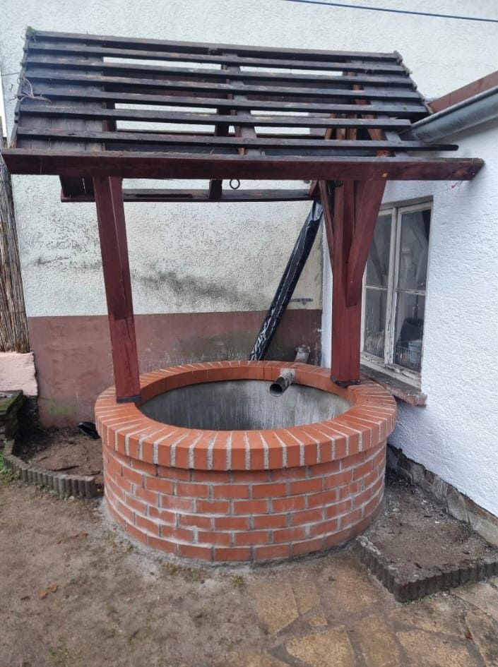 Ein gemauerter Brunnen mit Holzrahmen und -dach, neben einem weißen Gebäude.