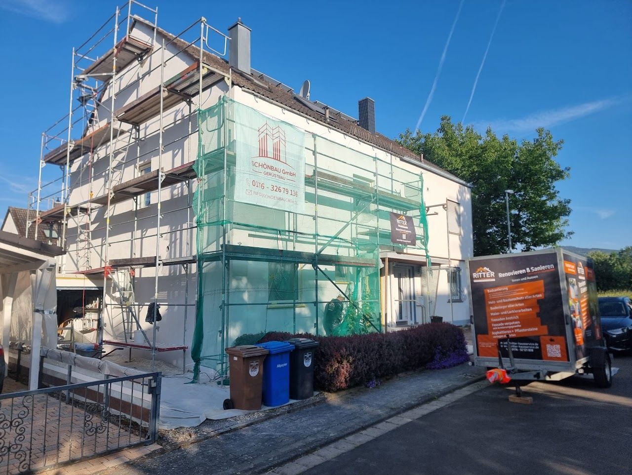 Die Hausfassade wird renoviert und ist mit Gerüst und Schutznetzen gesichert; ein Wohnwagen steht in der Nähe.
