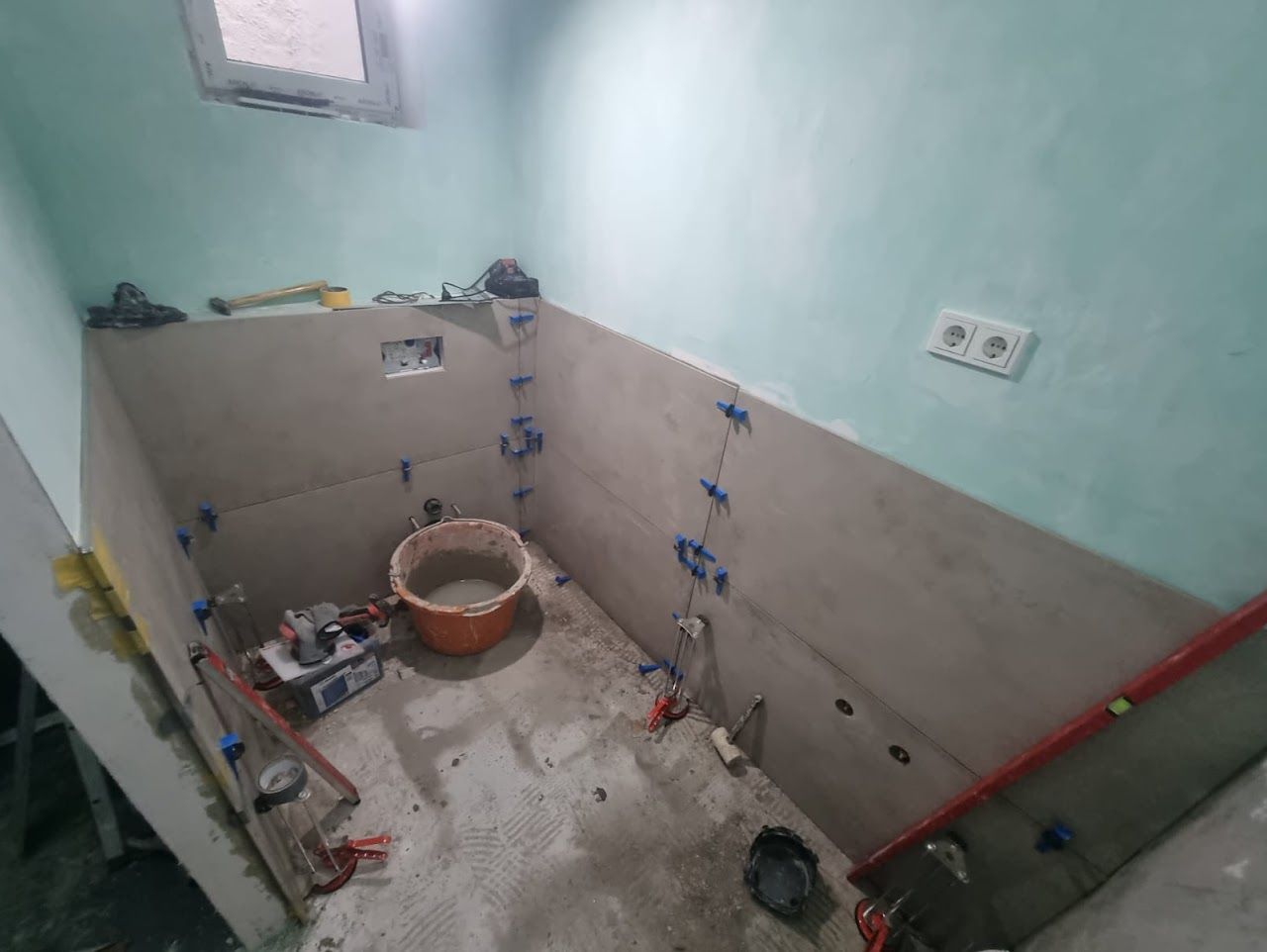 Badezimmer im Umbau, Wände teilweise gefliest; Werkzeug, Eimer und Wasserwaage sichtbar.