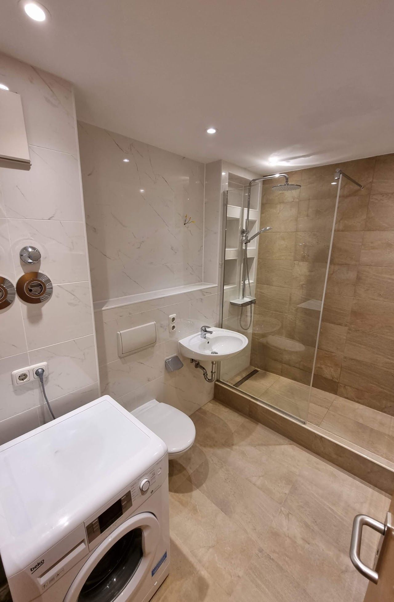 Ein kleines, modernes Badezimmer mit Dusche, Toilette, Waschbecken und Waschmaschine; hellbraune und beige Farbtöne.