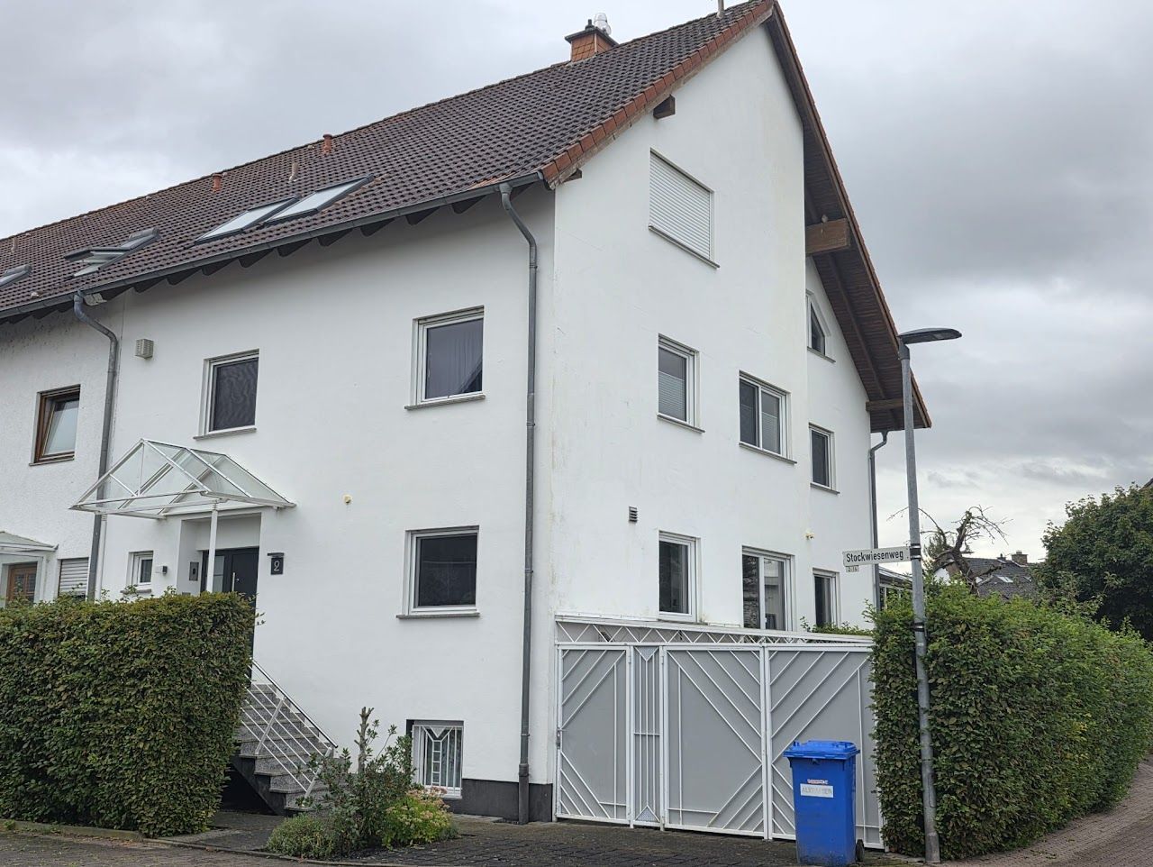 Weißes, mehrstöckiges Gebäude mit rotem Ziegeldach, roten Fenstern und einem blauen Mülleimer.