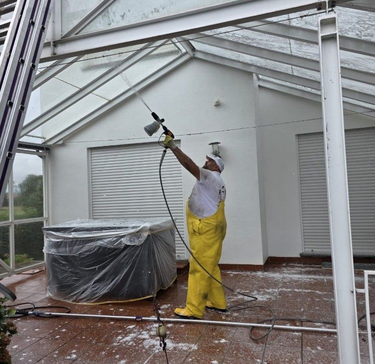 Mann in gelbem Schutzanzug reinigt mit einem Hochdruckreiniger eine Terrasse mit Glasdach.