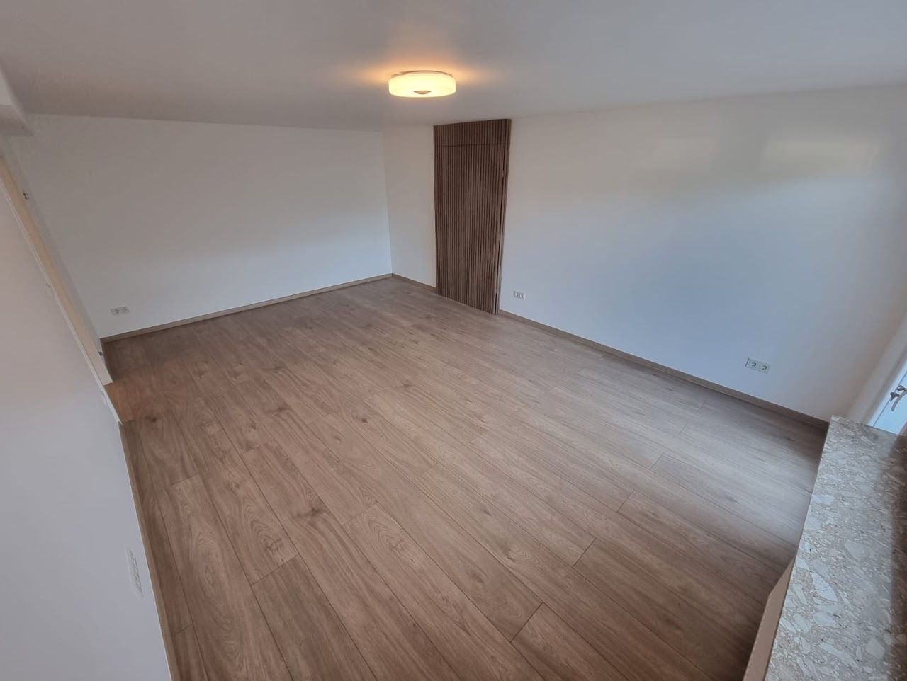 Leeres Zimmer mit Holzbodenoptik und einer Akzentwand aus Holz. Weiße Wände und eine Deckenleuchte.