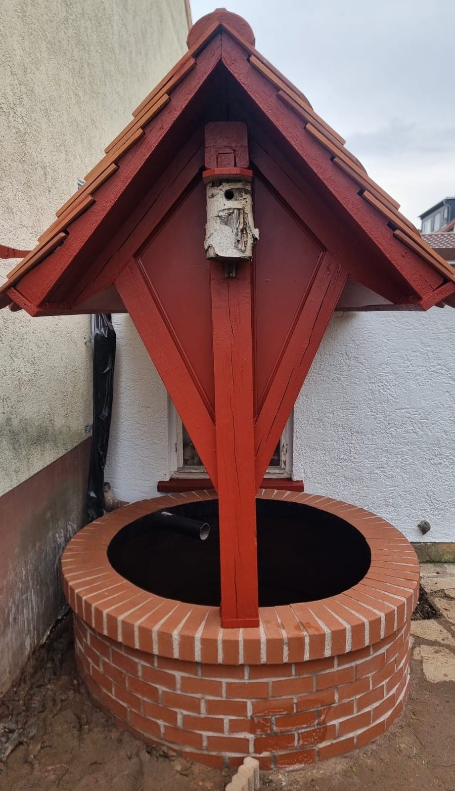 Ein rotgedeckter Wunschbrunnen mit gemauertem Sockel vor einer weiß-braunen Gebäudewand.