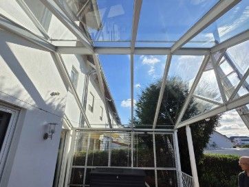 Terrasse mit Glasdach, transparenten Paneelen und weißem Metallrahmen, durch den man einen blauen Himmel sehen kann.