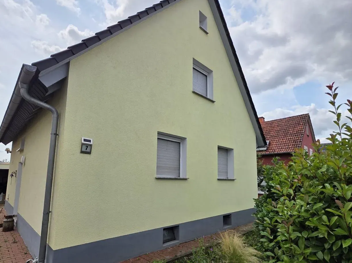 Kleines hellgelbes Haus mit grauen Zierelementen und Ziegeldach unter einem bewölkten Himmel