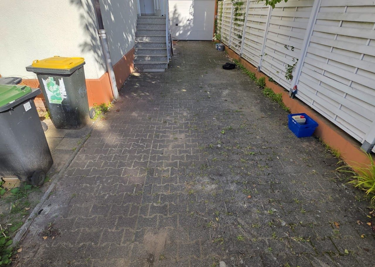 Schmaler gepflasterter Gehweg neben einem Gebäude und einem weißen Zaun, mit Mülltonnen und einem blauen Eimer.