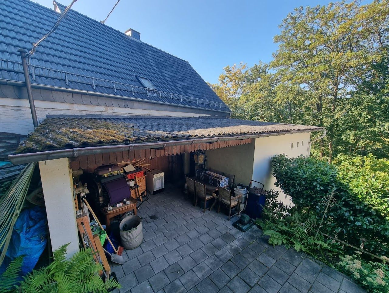 Überdachter Außenbereich mit vollgestellten Abstellräumen unter einem Dach. Bäume im Hintergrund.