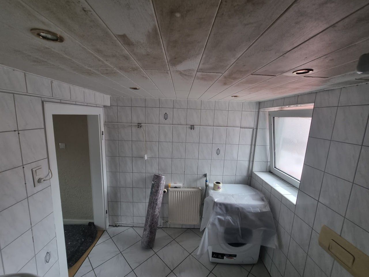 Badezimmer mit weißen Fliesenwänden, Deckenpaneelen und einer mit Plastikfolie abgedeckten Waschmaschine.