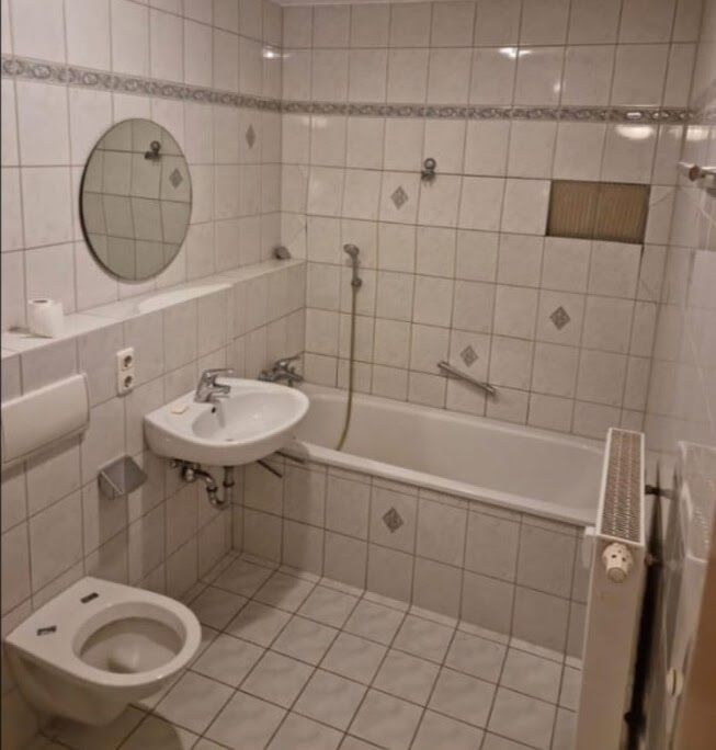 Kleines, weiß gefliestes Badezimmer mit Toilette, Waschbecken, Badewanne und rundem Spiegel.