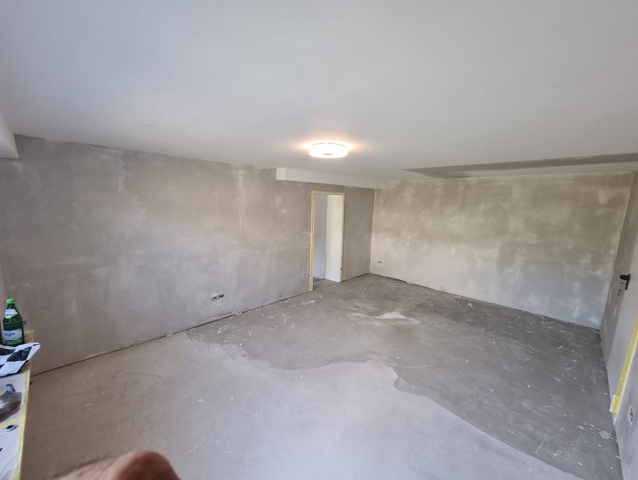 Leerer Raum mit grauen Betonwänden und -boden, einzelner Deckenleuchte, Türöffnung sichtbar.