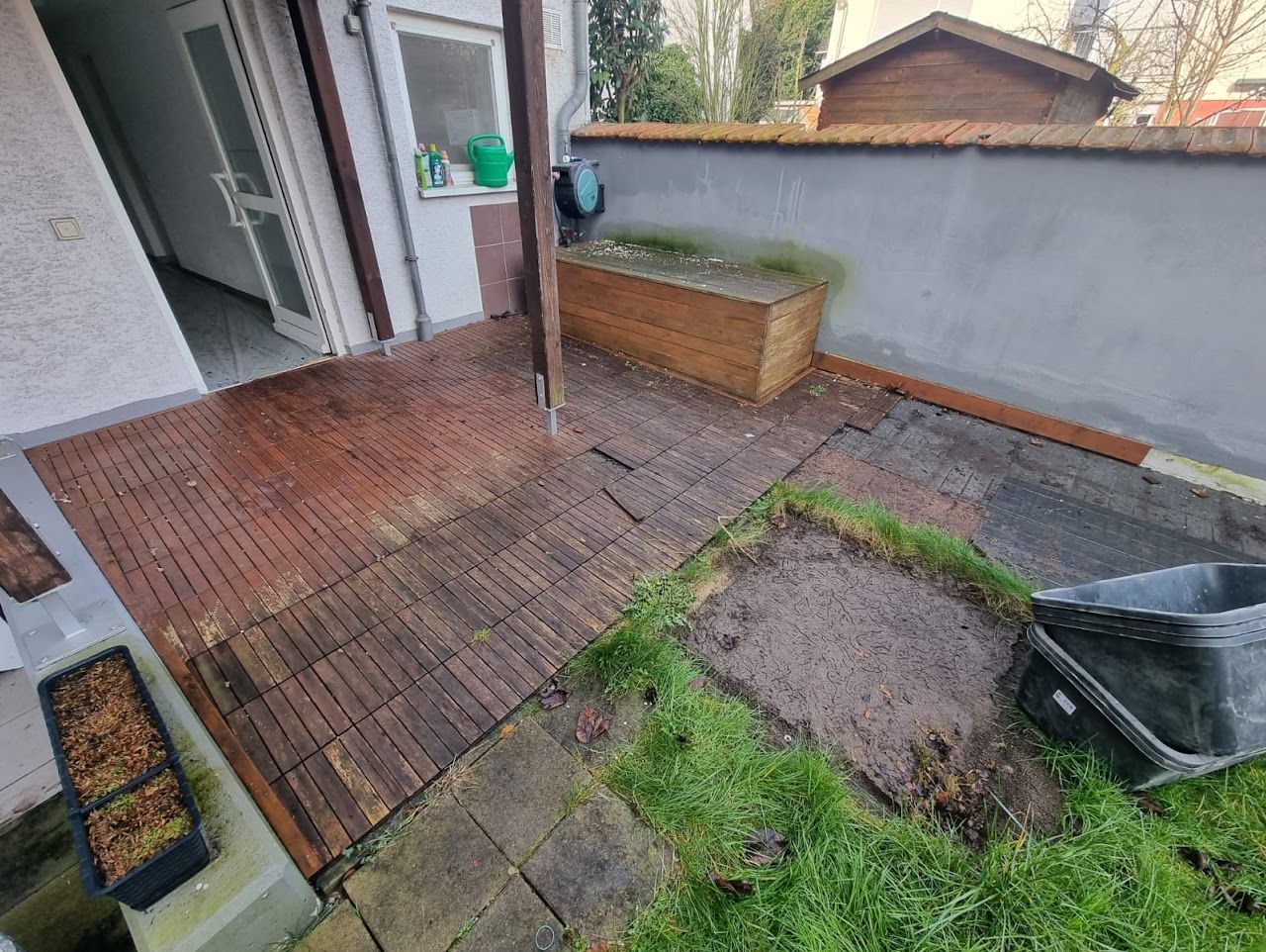 Eine Holzterrasse mit Pflanzkübel, Teil eines Gartens und einer Tür zu einem Gebäude.