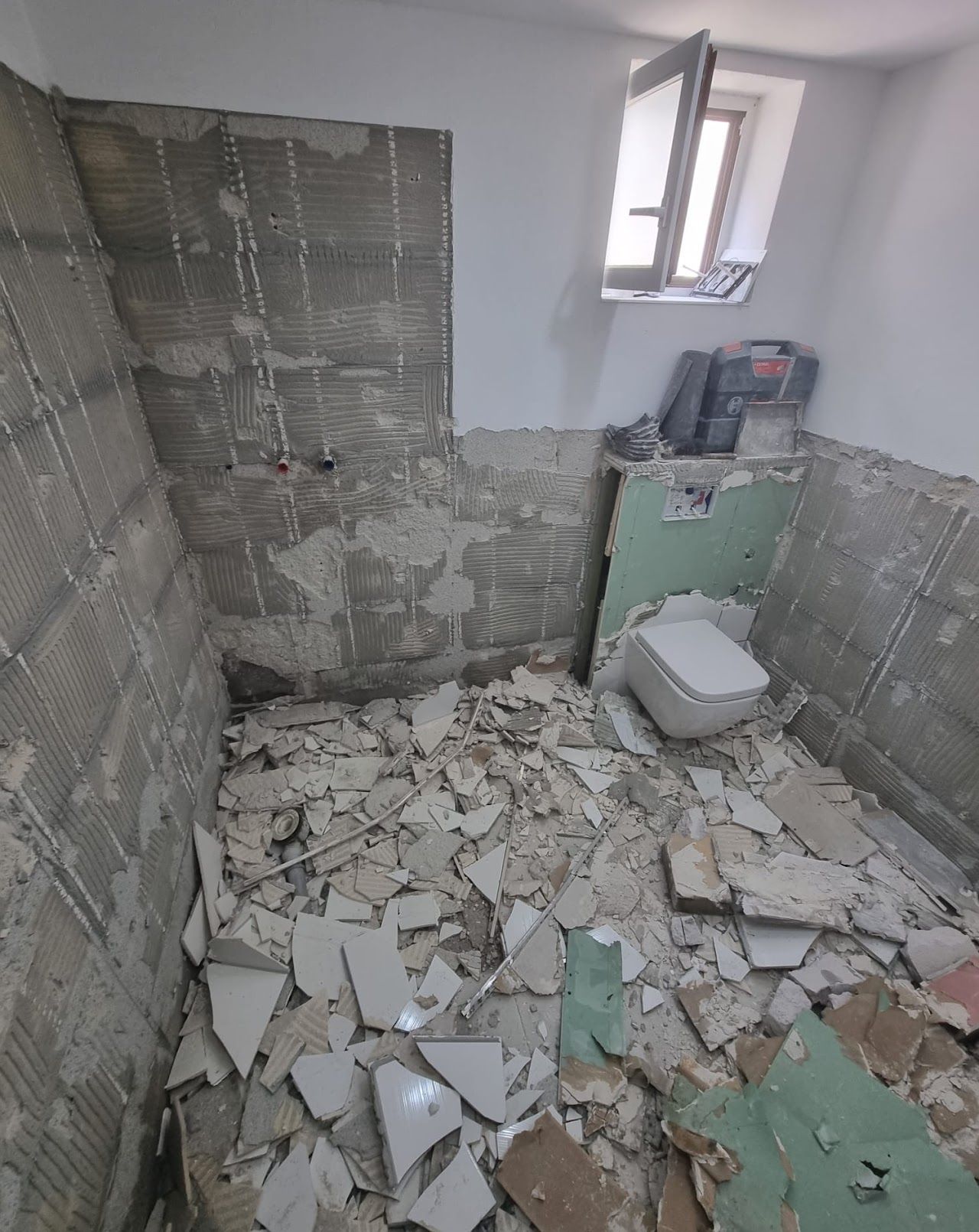 Badezimmerrenovierung im Gange; Fliesen und Bauschutt auf dem Boden, freiliegende Wände, Toilette vorhanden, kleines Fenster.