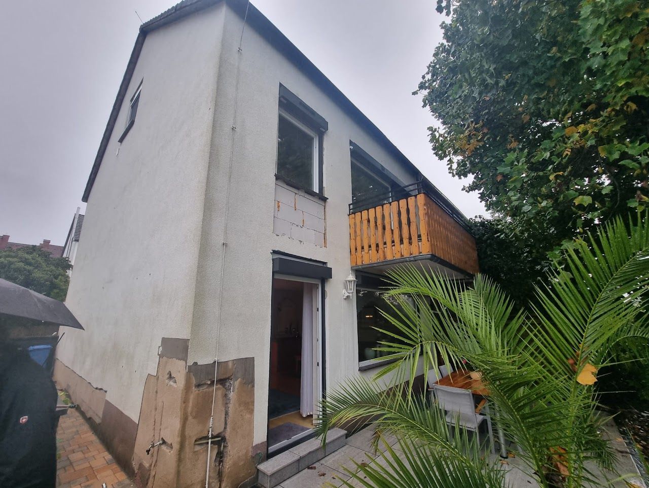 Zweistöckiges weißes Haus mit Holzbalkon, sichtbarer Tür und Palme davor, an einem bewölkten Tag.