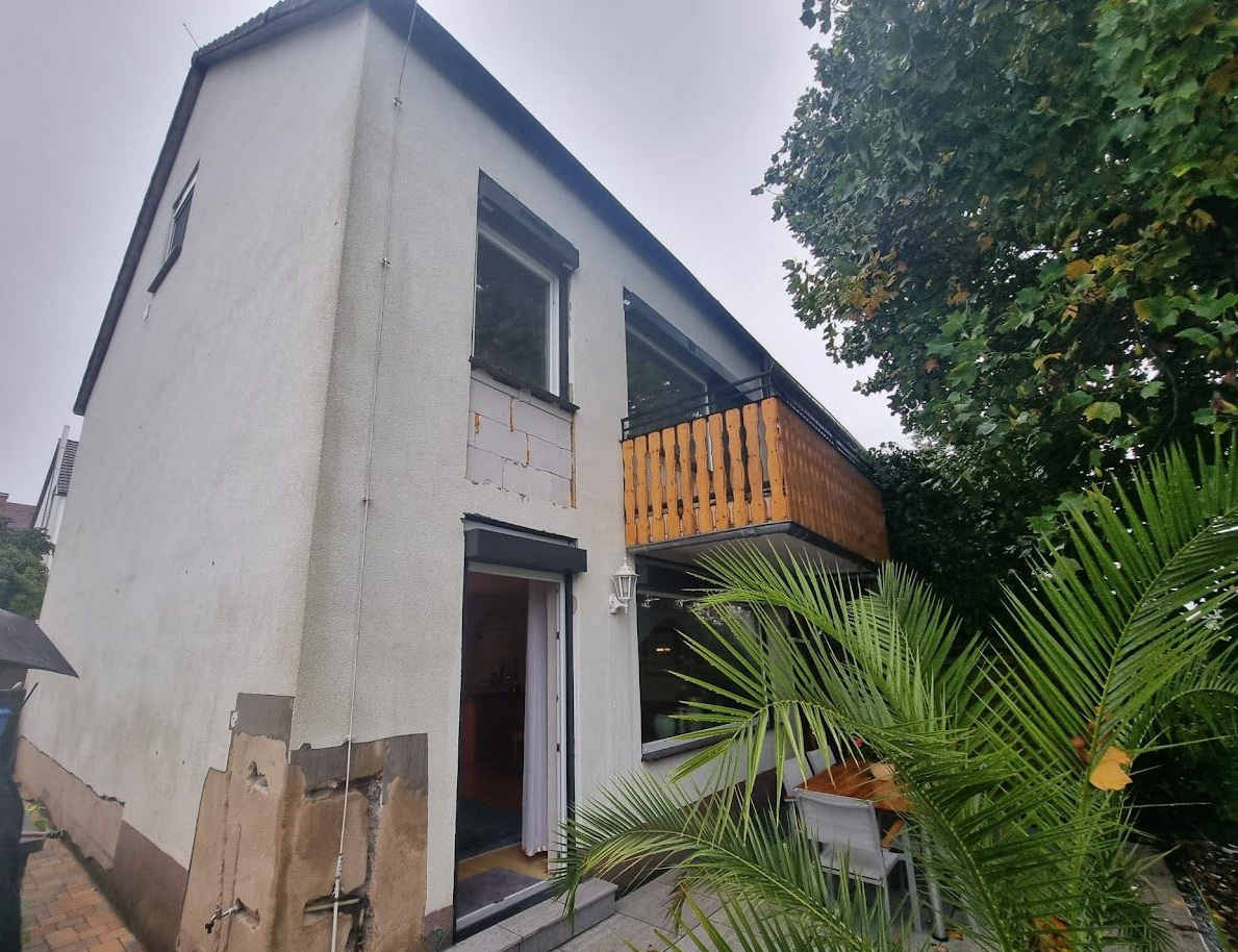 Außenansicht eines schmalen, weißen, zweistöckigen Hauses mit Holzbalkon und Gartenpflanzen davor