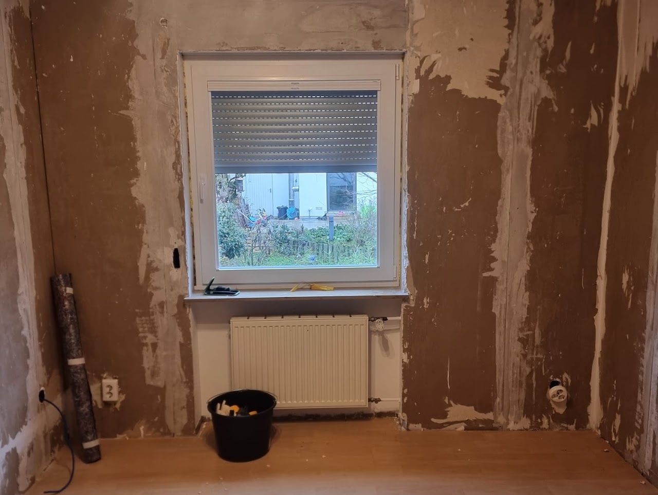 Innenansicht eines Zimmers während der Renovierung, mit Fenster, Heizkörper, freiliegenden Wänden und Werkzeugen.