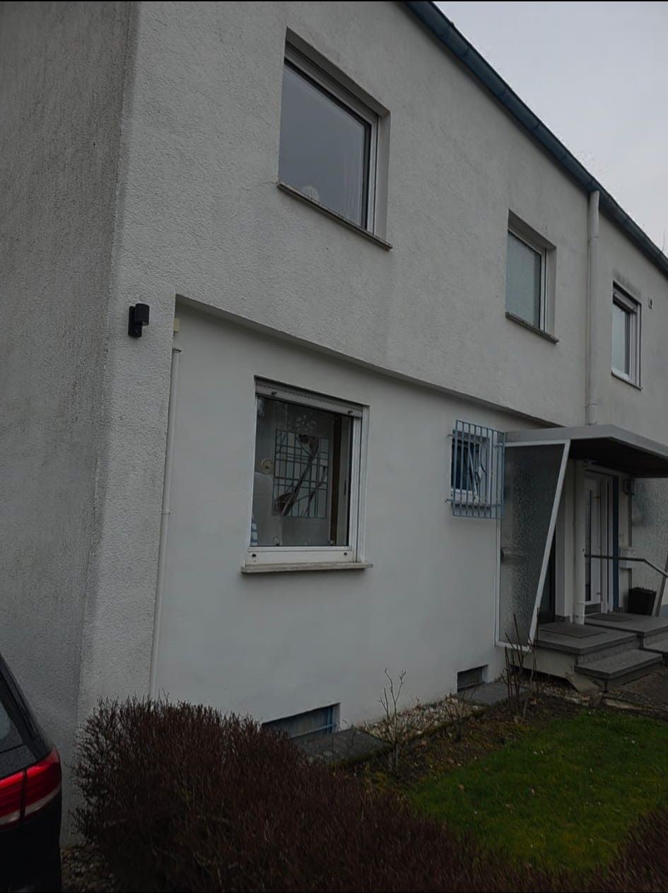 Weißes, zweistöckiges Gebäude mit Fenstern. Im Vordergrund ein dunkles Auto. Bewölkter Himmel.