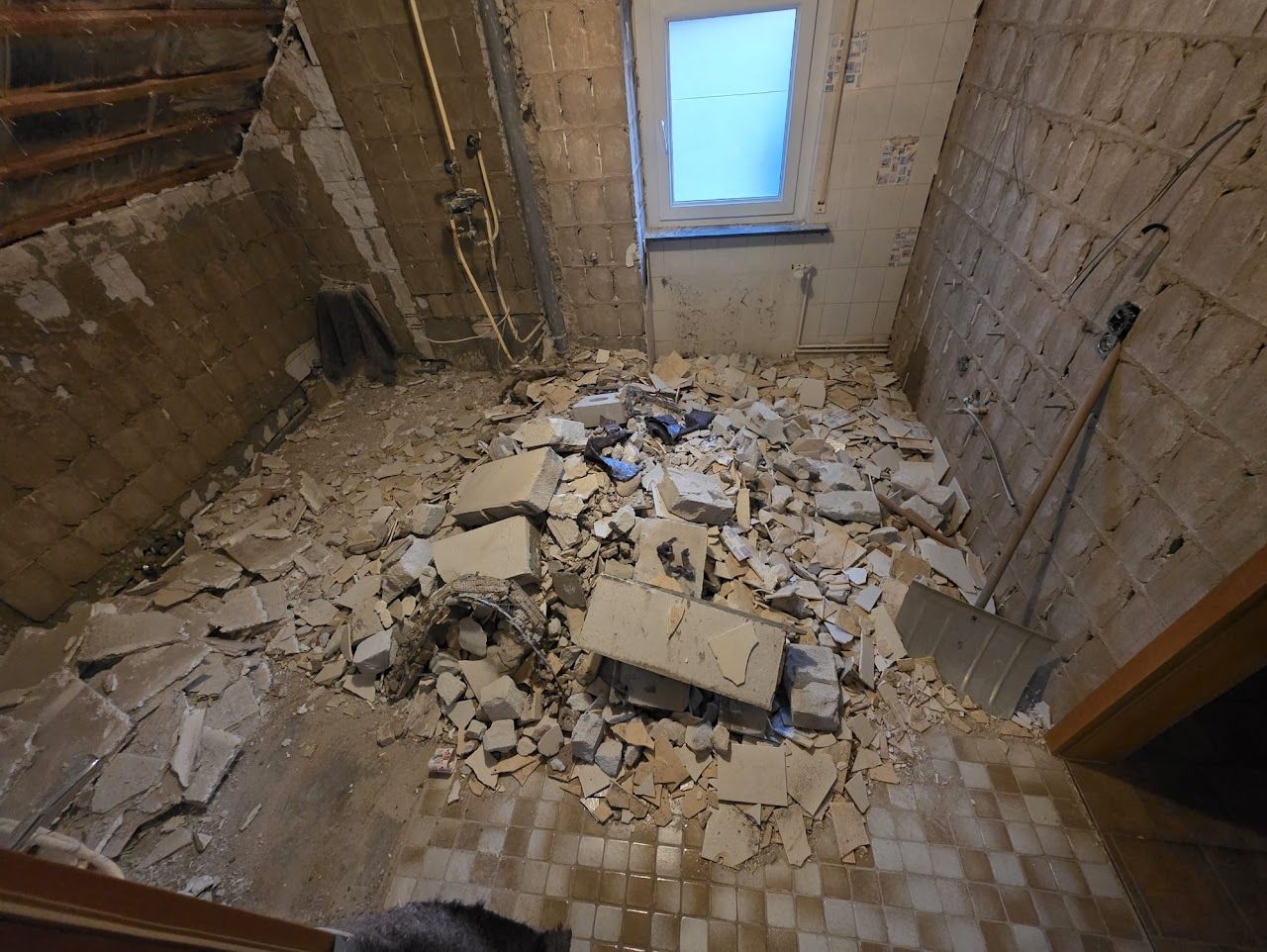 Zimmer im Umbau, Bauschutt auf dem Boden, Fenster darüber. Sichtbare Balken, kahle Wände, neutrale Farbtöne.