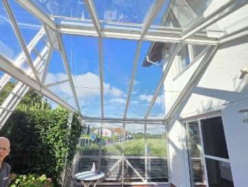 Wintergarten mit Glasdach und Blick ins Grüne; in der linken Ecke ist eine Person teilweise zu erkennen.