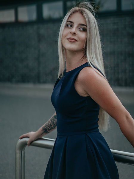 Blonde Frau in einem dunkelblauen Kleid lehnt an einem Geländer und blickt mit einem leichten Lächeln nach oben. Im Freien.