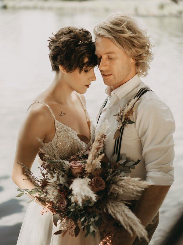 Brautpaar umarmt sich; Braut hält Blumenstrauß, Bräutigam trägt Hosenträger, in der Nähe von Wasser.