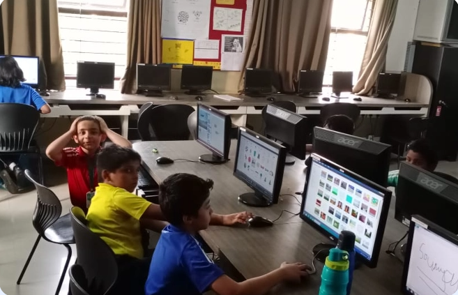A group of children are sitting in front of computer monitors