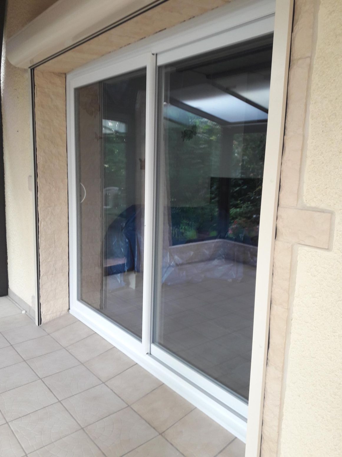 Porte-fenêtre coulissante à cadre blanc donnant sur une terrasse carrelée et un mur beige.