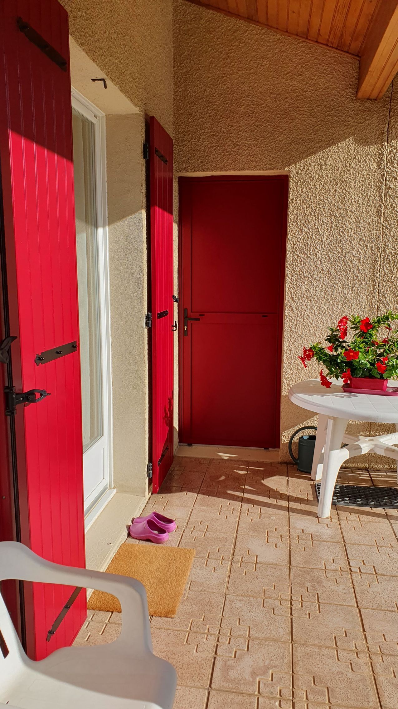 Une terrasse ensoleillée avec un sol carrelé, des volets rouges, une table blanche ornée de fleurs rouges et une chaise au premier plan.