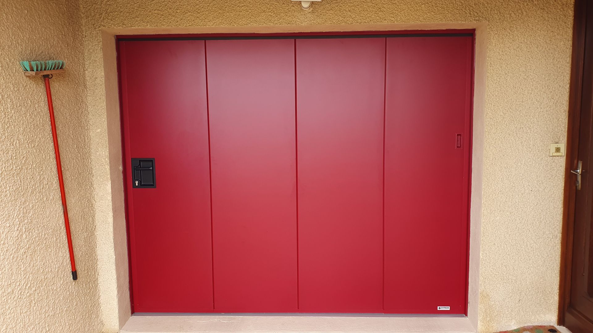 Une porte de garage lisse, rouge massif, à quatre panneaux, montée sur un mur en stuc beige, avec une poignée noire sur le côté gauche.