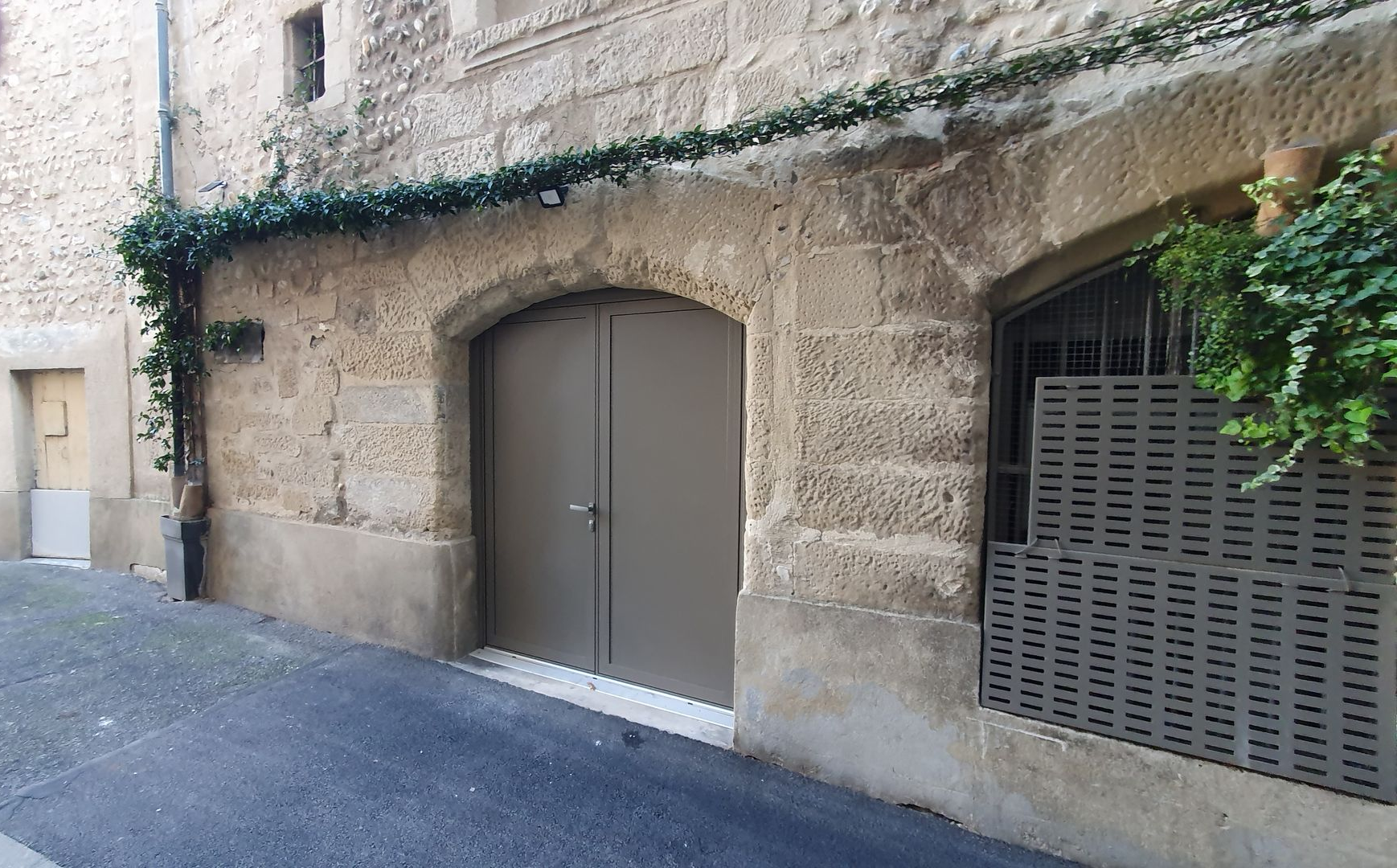 Un bâtiment en pierre avec une double porte grise et une fenêtre adjacente munie d'une grille métallique, encadré par des vignes grimpantes, donnant sur une rue.