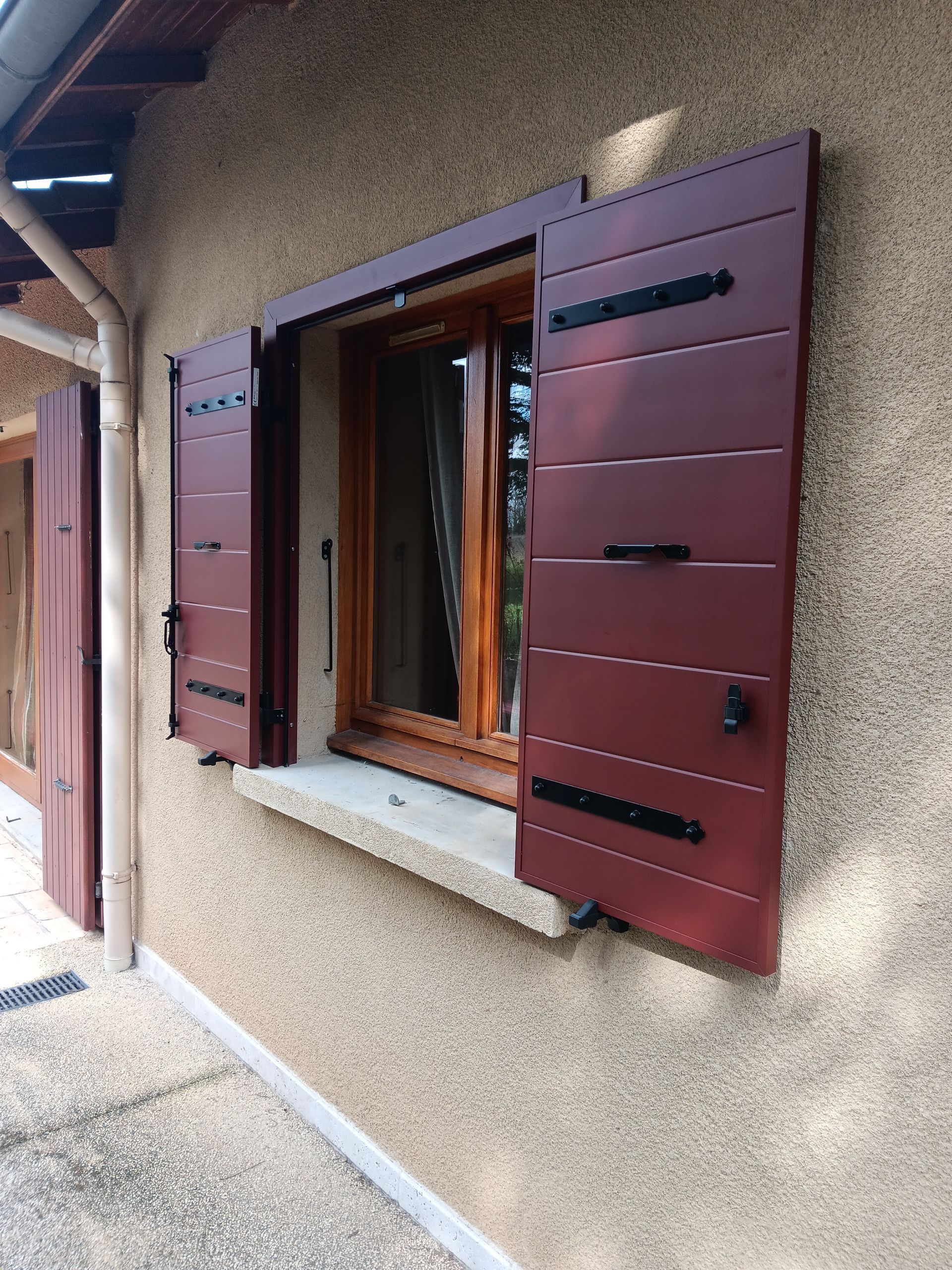 Une fenêtre en bois aux volets ouverts, rouge foncé, à planches horizontales, se détache sur un mur en stuc texturé beige.