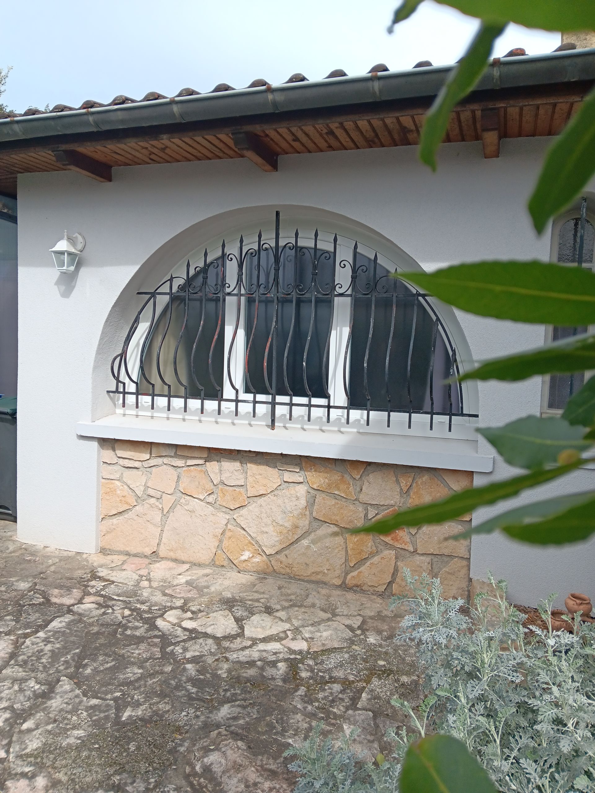Un bâtiment blanc avec une fenêtre cintrée protégée par une grille décorative en métal noir, situé au-dessus d'un mur carrelé en pierre.
