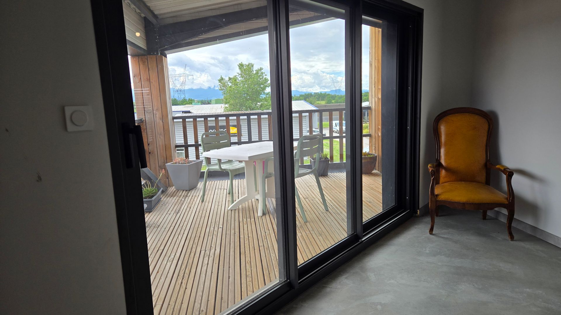 Pièce intérieure avec portes coulissantes ouvertes donnant sur un balcon, avec vue sur les arbres et l'eau, et une chaise en bois contre le mur.