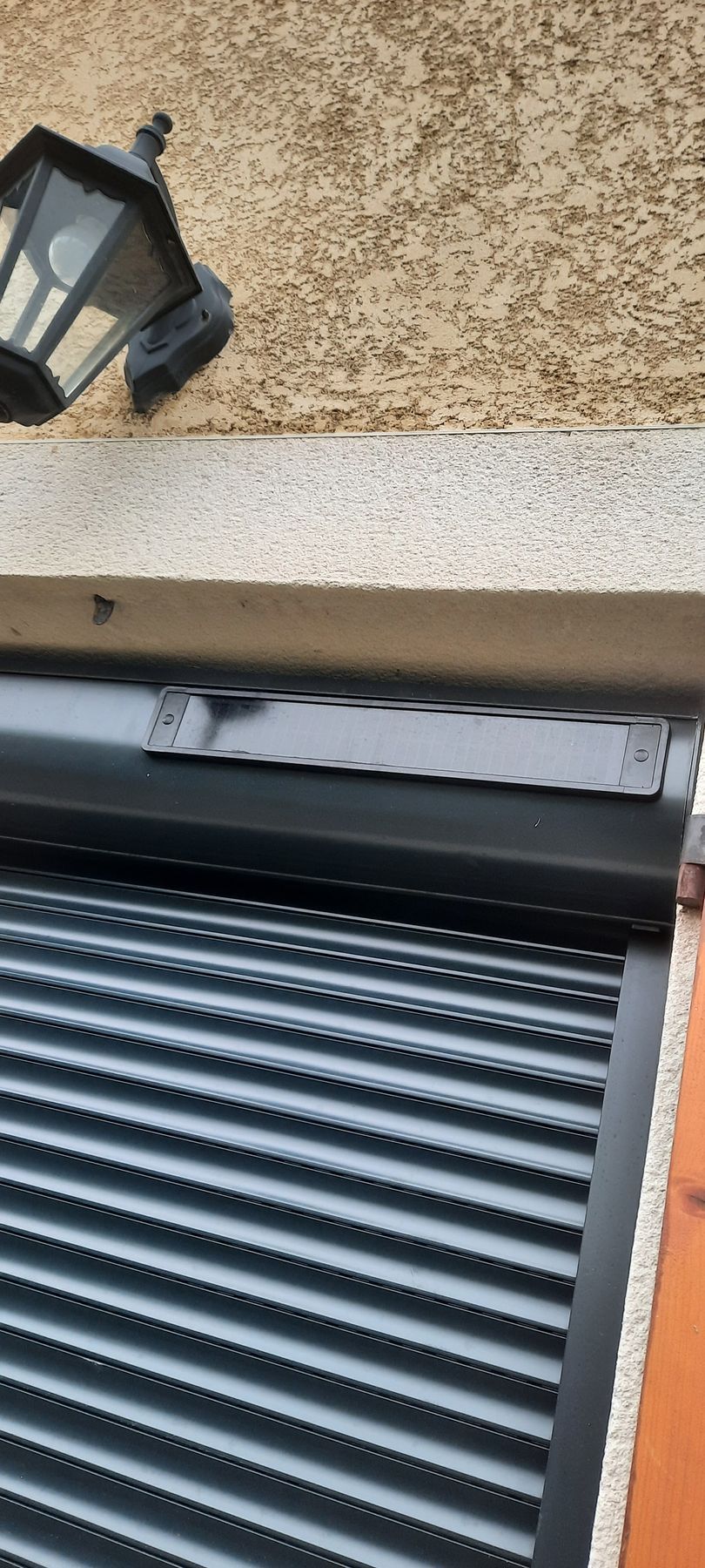 Vue de dessus d'un volet roulant gris foncé fermé sur une fenêtre, monté sous un mur en pierre texturée et un éclairage extérieur.