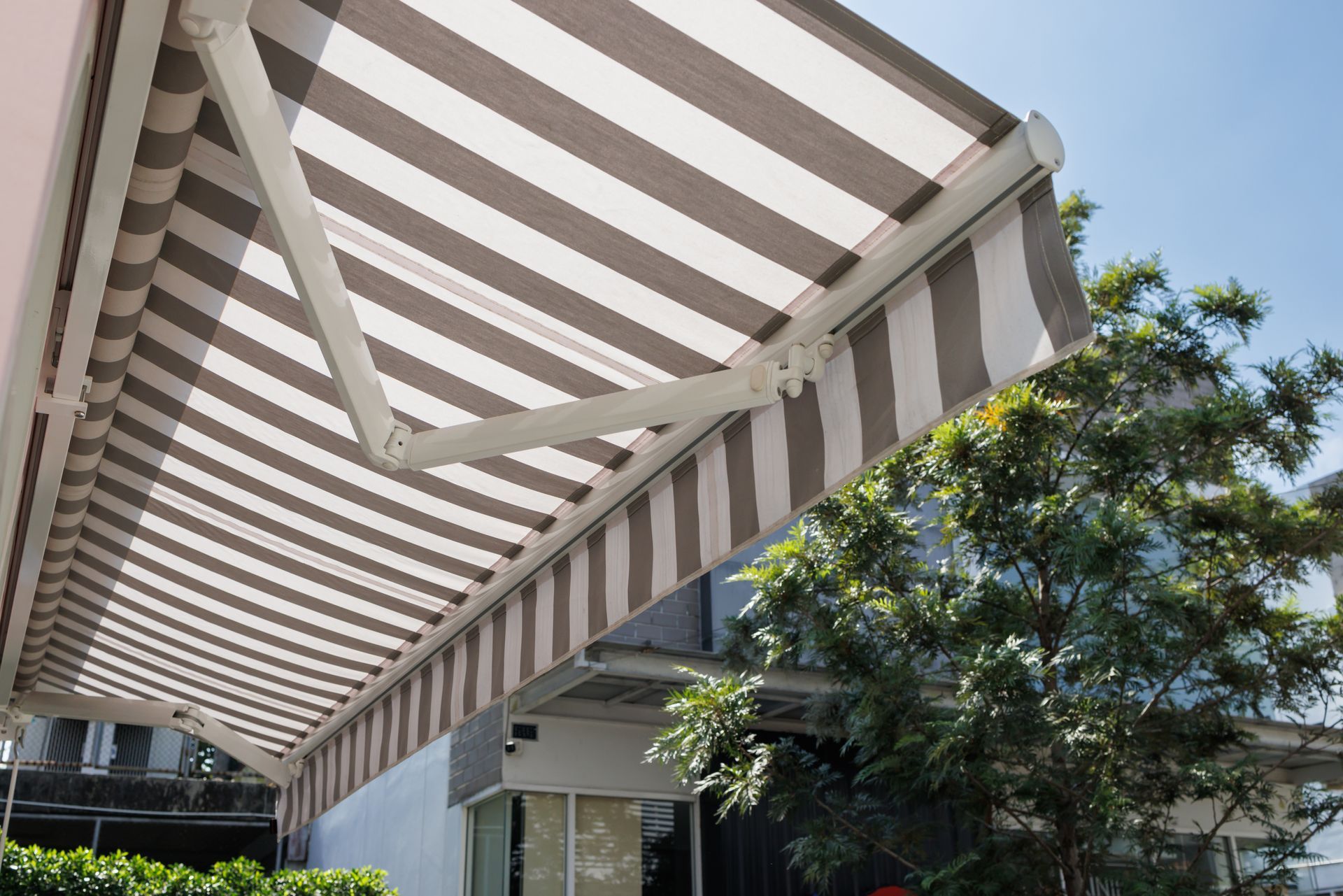 Un store rétractable à rayures brunes et blanches s'étendait au-dessus d'un espace extérieur avec des arbres et un bâtiment en arrière-plan.