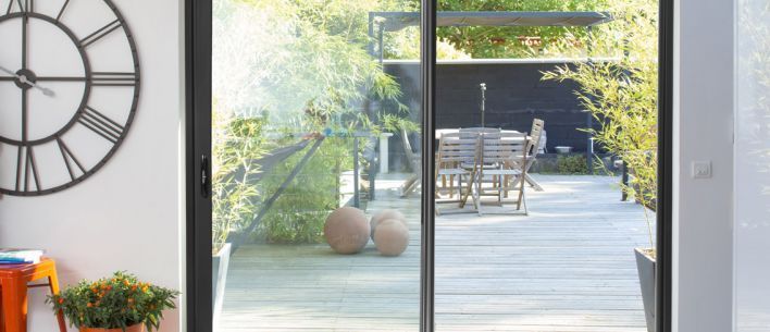 Vue à travers une porte coulissante en verre sur un patio ensoleillé avec un ensemble de salle à manger et des plantes luxuriantes, à côté d'une horloge murale.