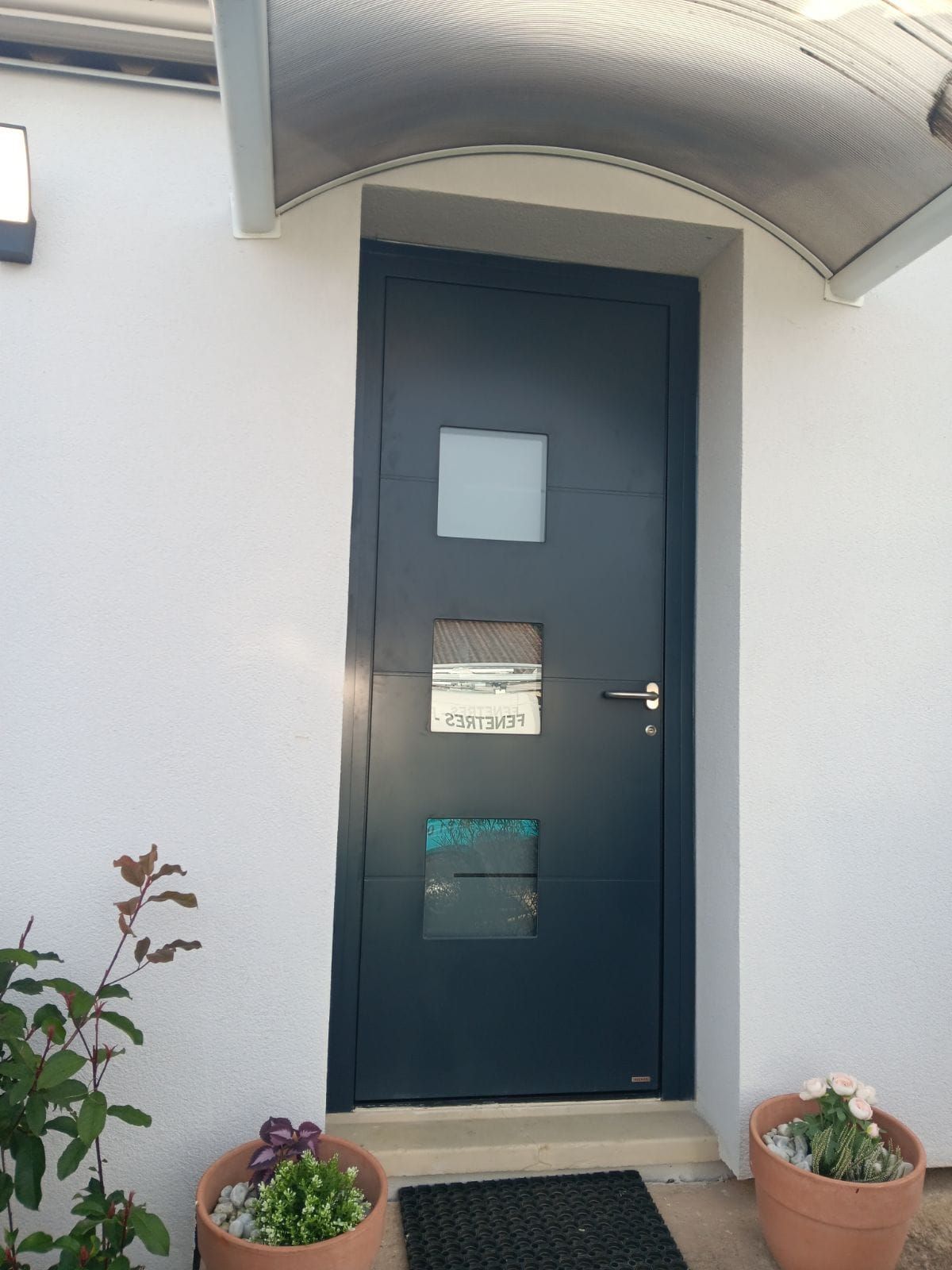 Une porte d'entrée gris foncé avec trois fenêtres carrées en verre, encastrée dans un mur blanc avec des plantes en pot de chaque côté.