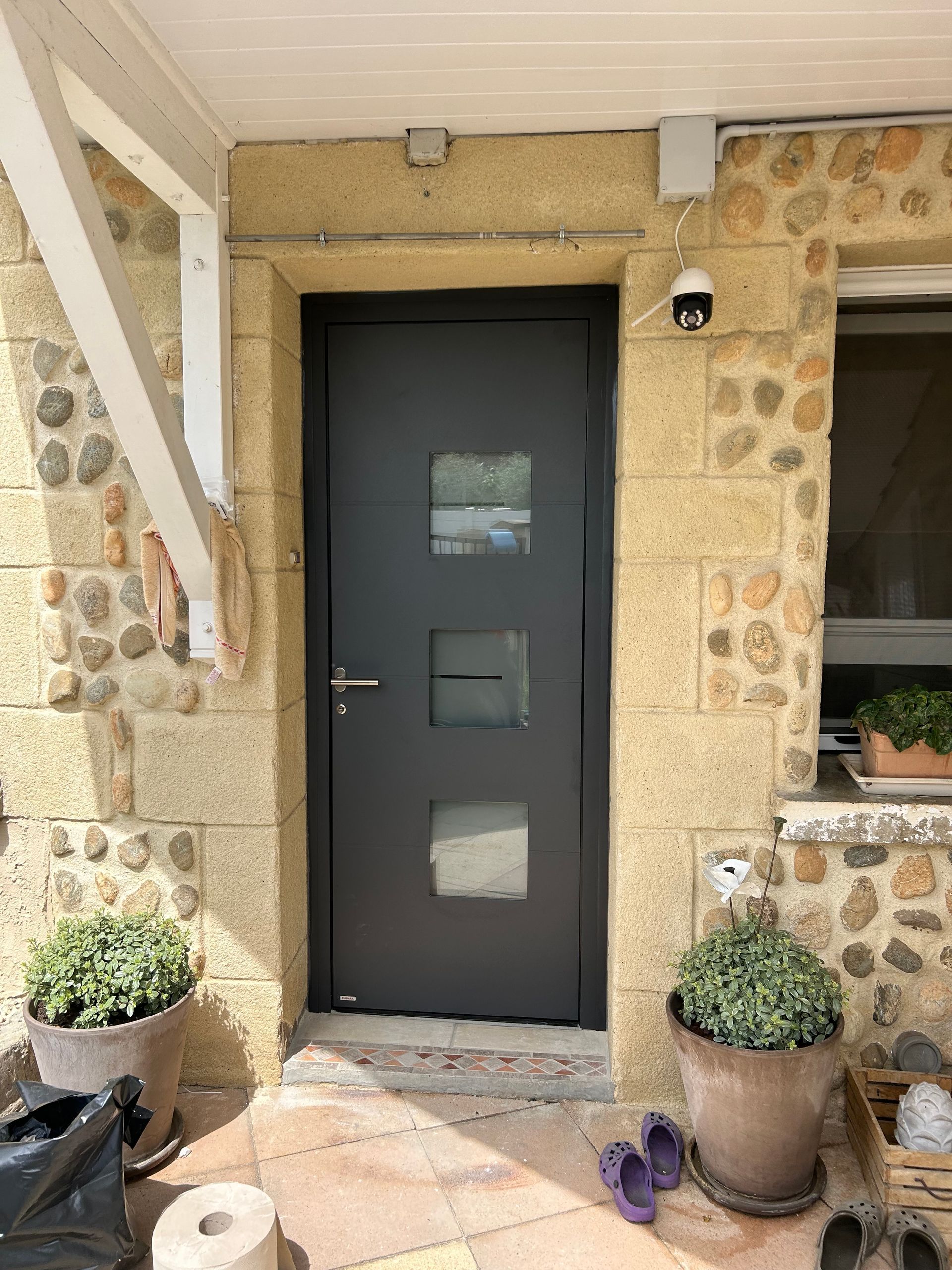 Entrée principale avec une porte sombre, des murs en pierre, des plantes en pot et une petite fenêtre sur la droite.