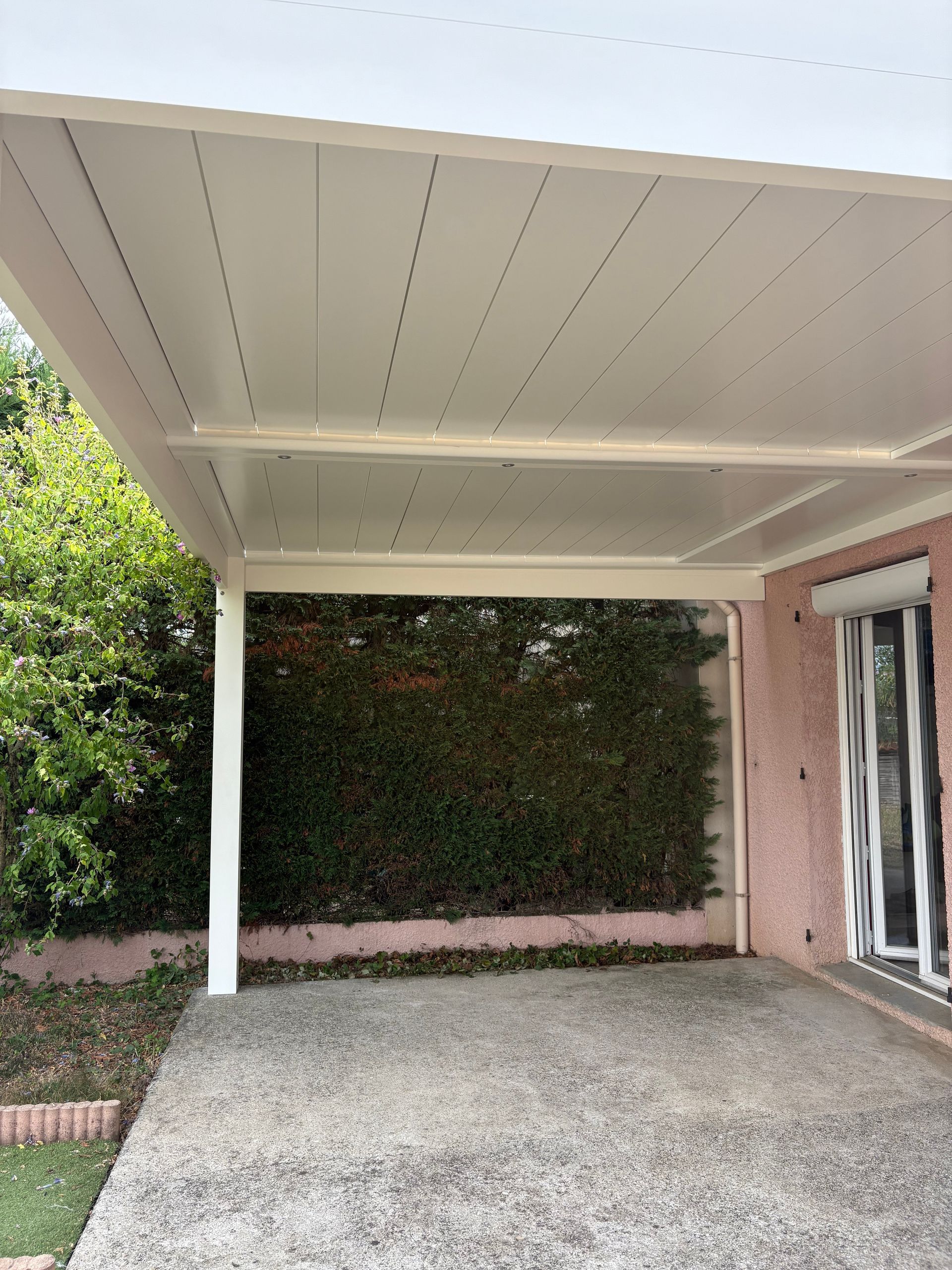 Une terrasse couverte en béton jouxte une maison rose avec une haie verte en arrière-plan.
