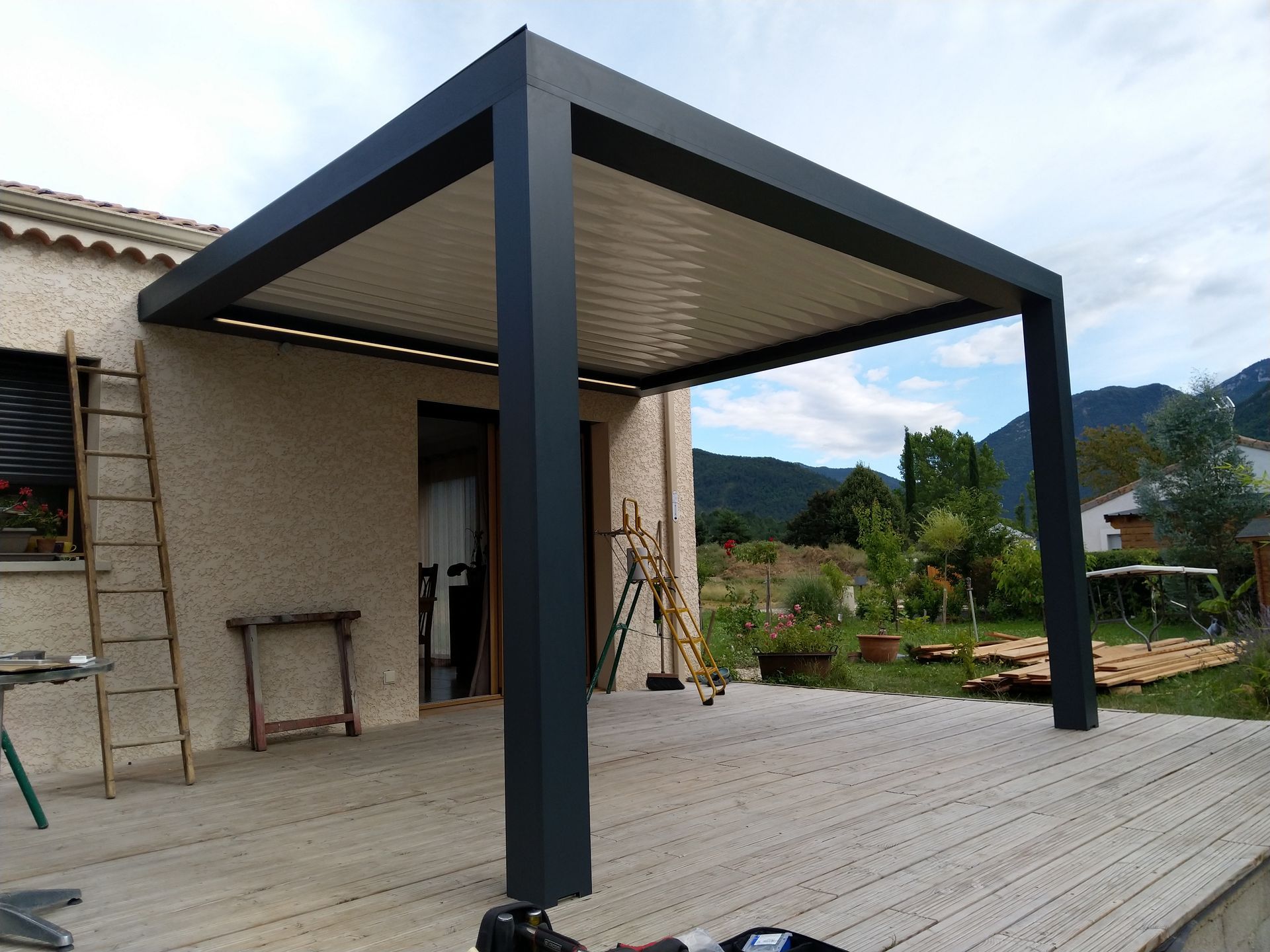 Une pergola moderne à persiennes gris foncé, adossée à une maison beige, surplombe une terrasse en bois offrant une vue sur la montagne.