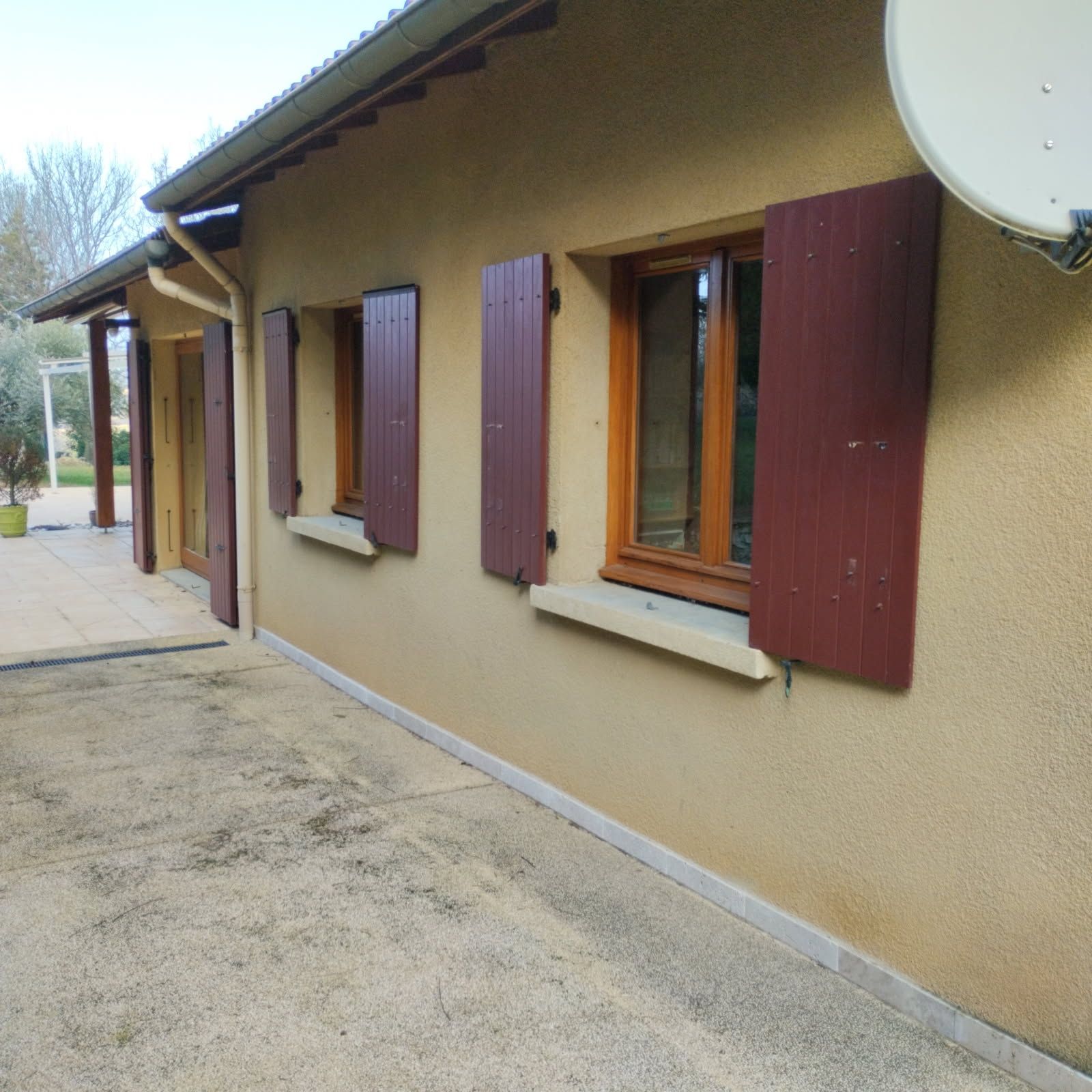 Vue latérale d'une maison de plain-pied de couleur beige, avec des volets en bois brun et une antenne parabolique fixée sur le mur extérieur.
