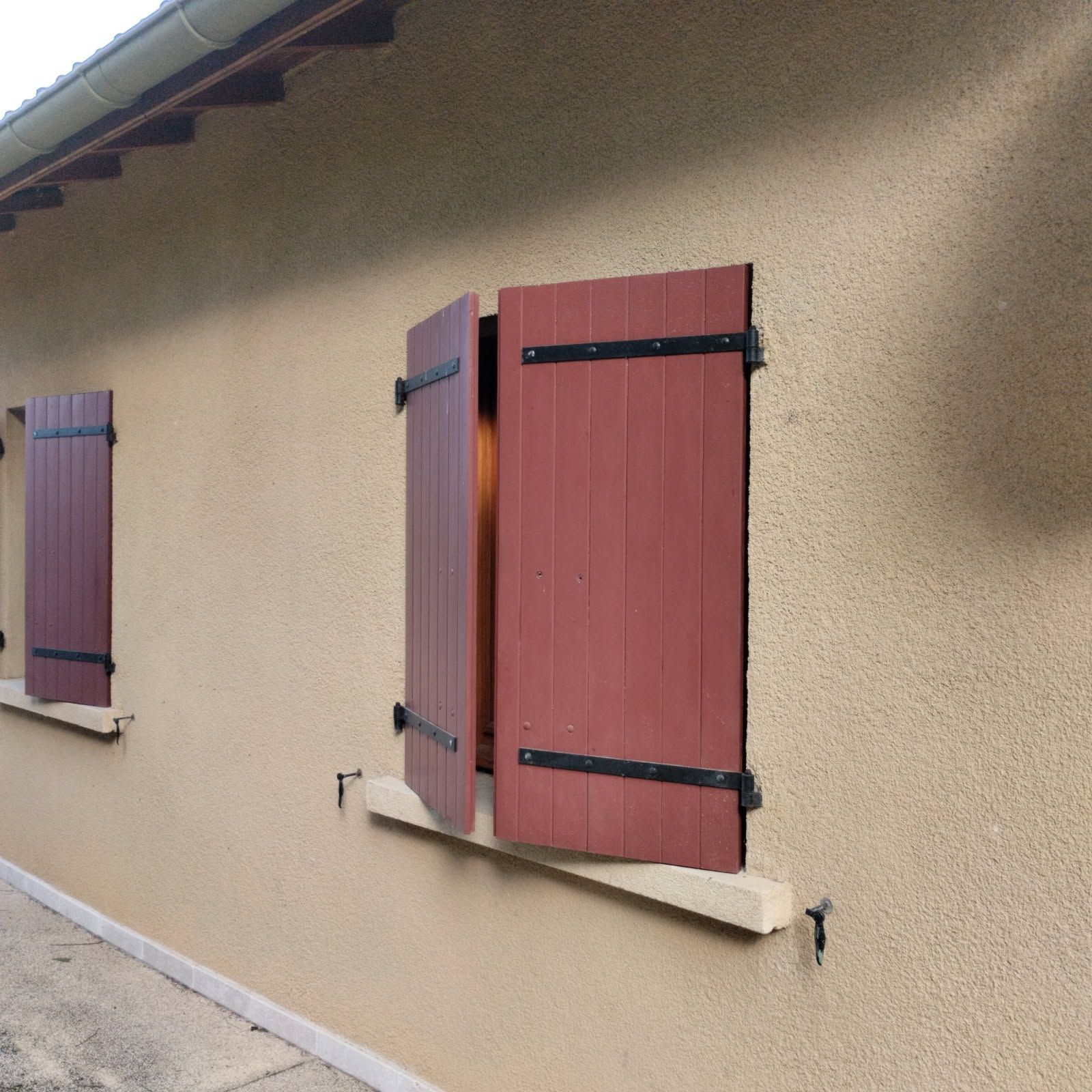 Volets en bois rouge sur deux fenêtres, devant un mur extérieur texturé, brun clair, d'une maison.