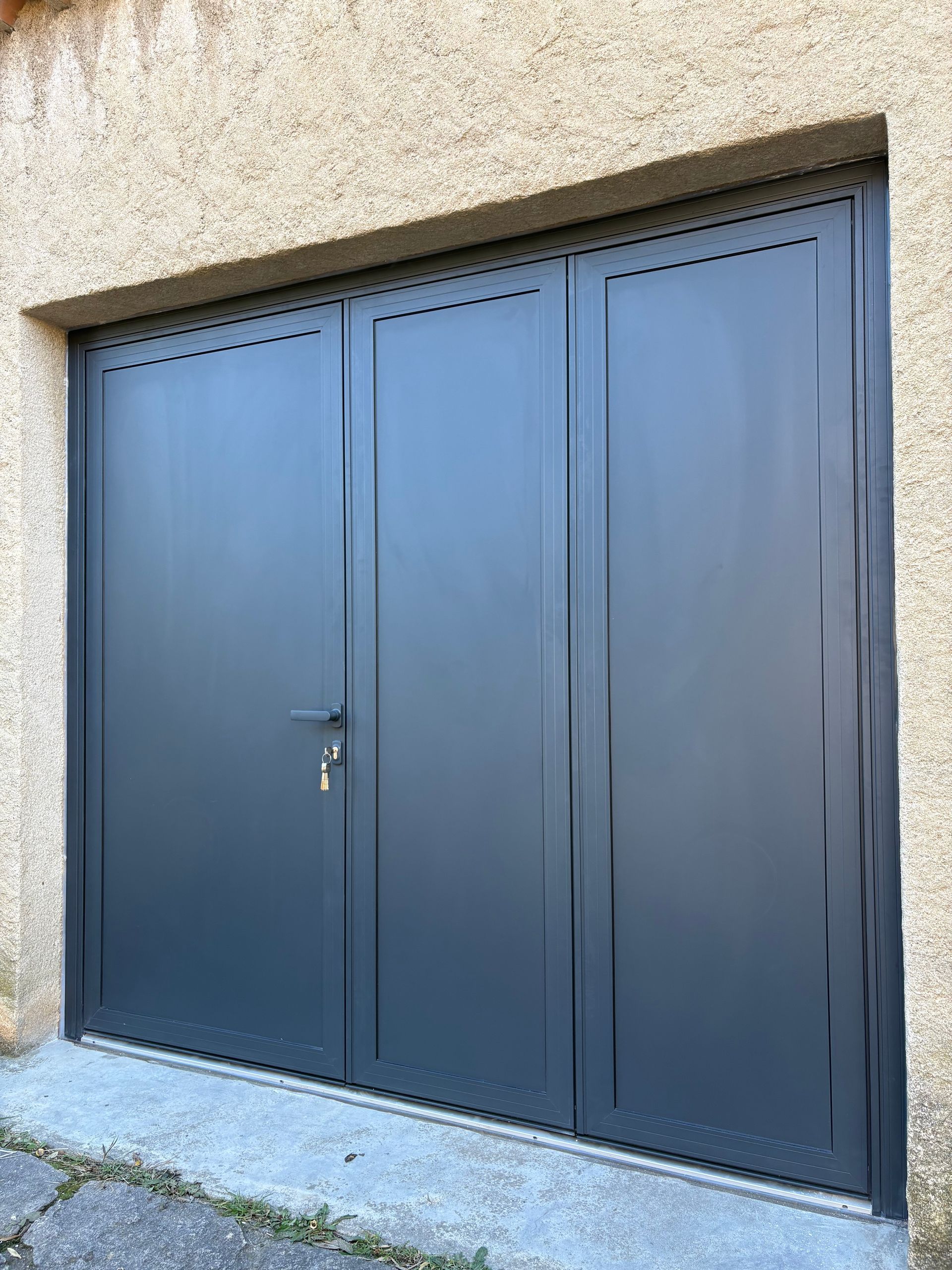 Porte de garage double en métal noir encastrée dans un mur en stuc beige, avec une petite poignée latérale et une serrure.